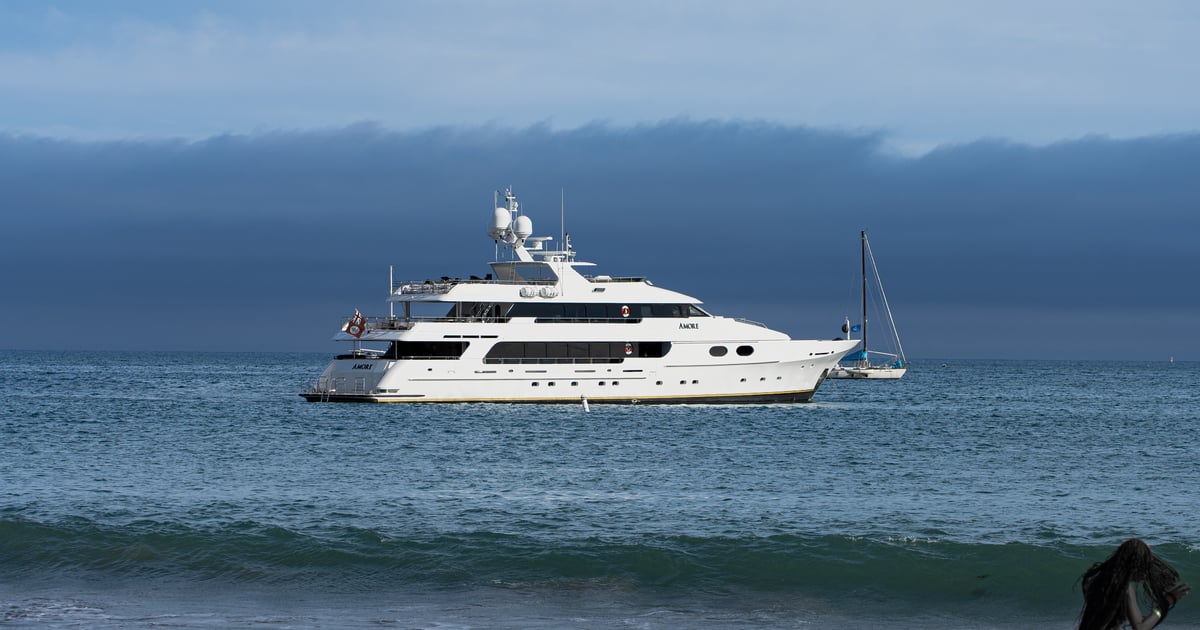 White yacht anchored offshore beneath a dark wall of summer fog